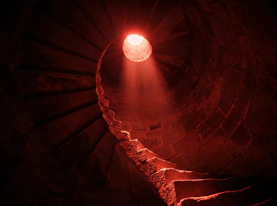 A spiral stone staircase lit by a shaft of red light from an oculus above.